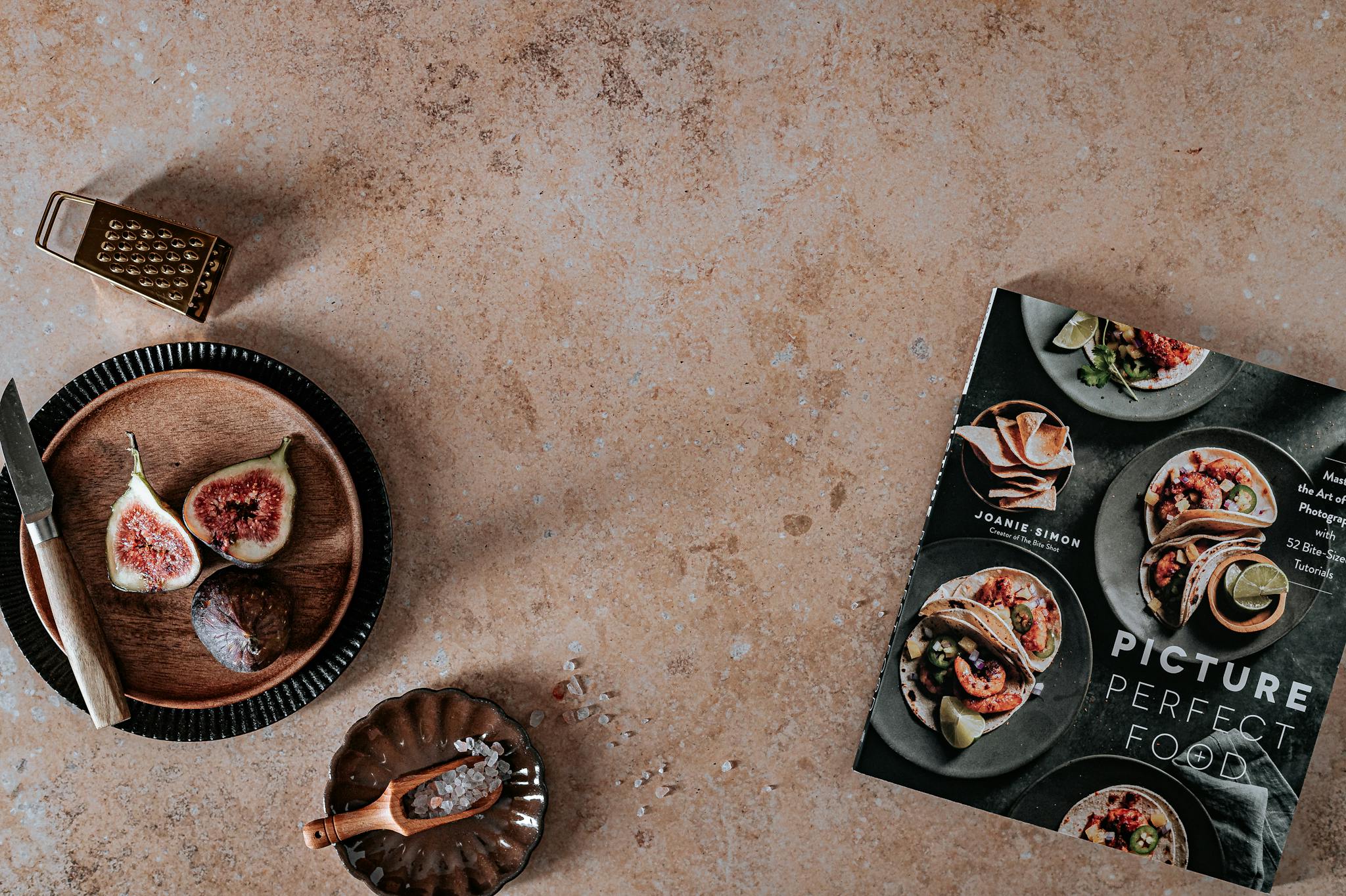A flat lay shot featuring figs, a knife, and a cookbook on a rustic surface.